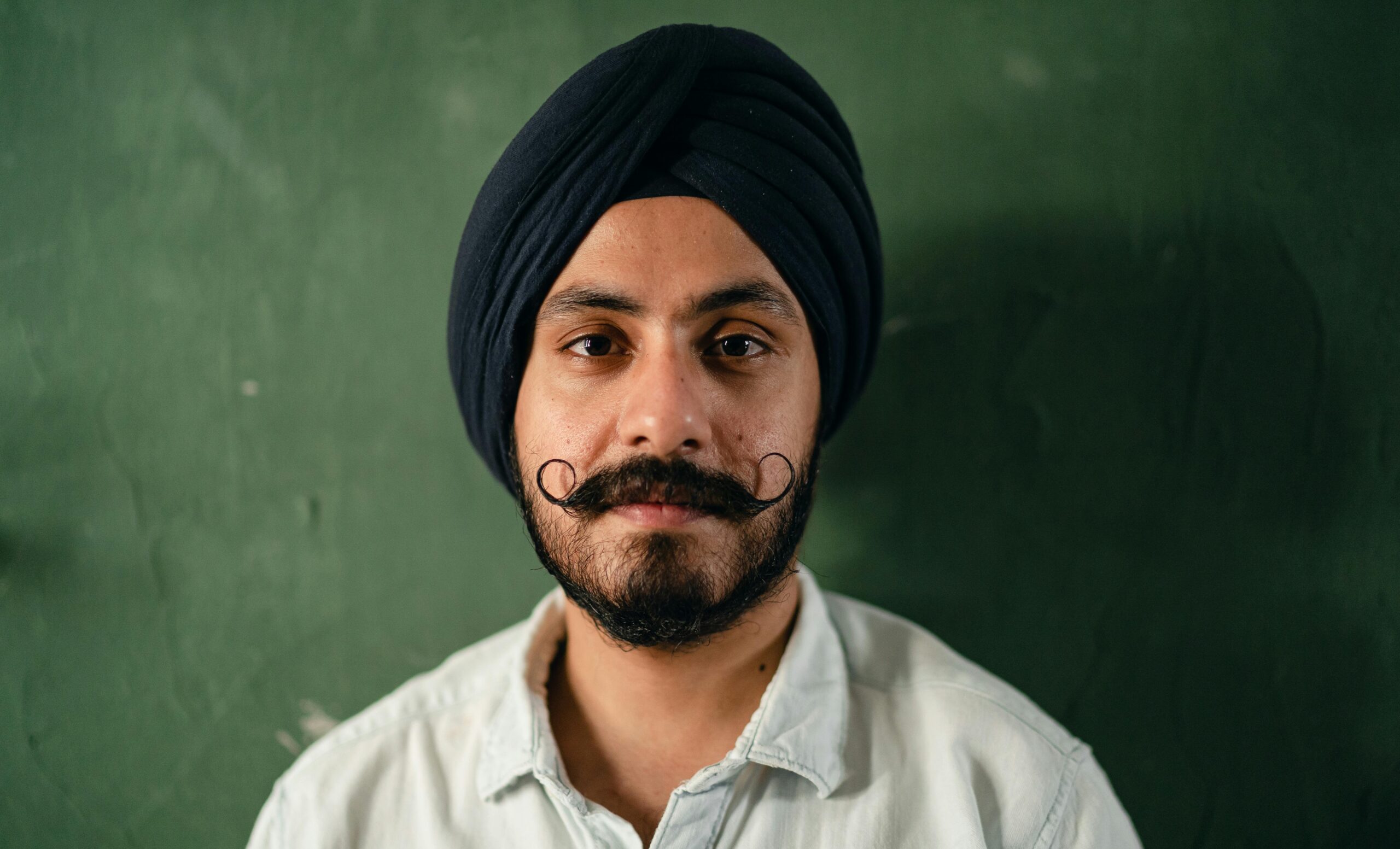 Portrait of content relaxed Sikh male in casual shirt and wearing black traditional turban and handlebar mustache standing against green scratched wall and looking at camera