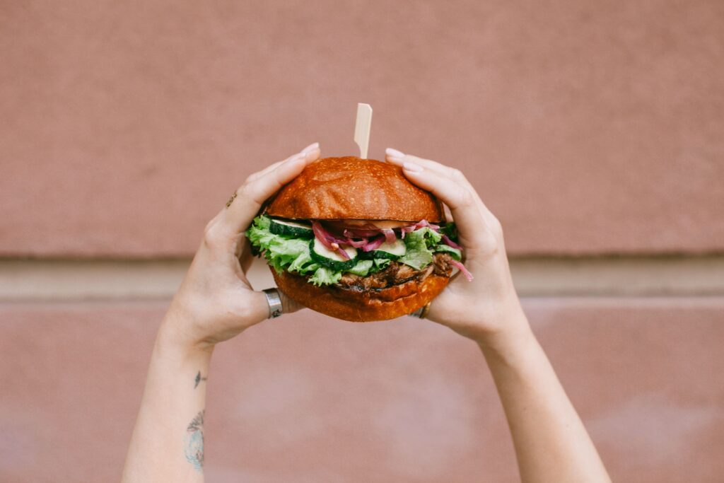 A delicious gourmet burger with fresh greens held by tattooed hands outdoors.
