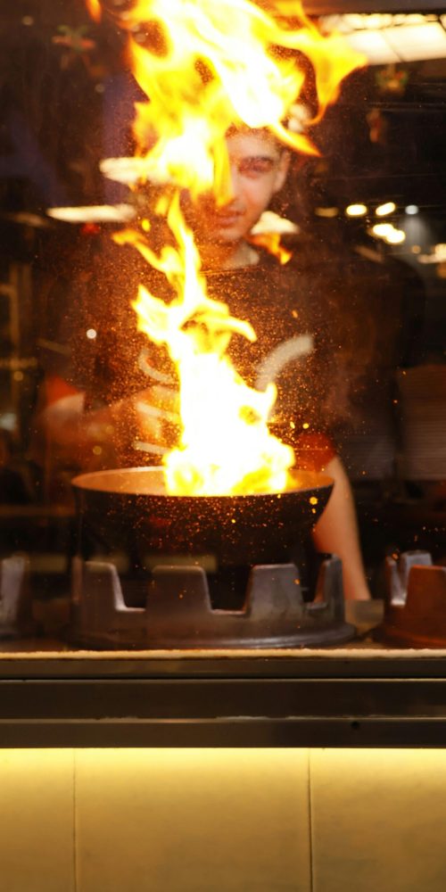 A chef creates a dramatic flare-up while cooking with a pan in a bustling kitchen.